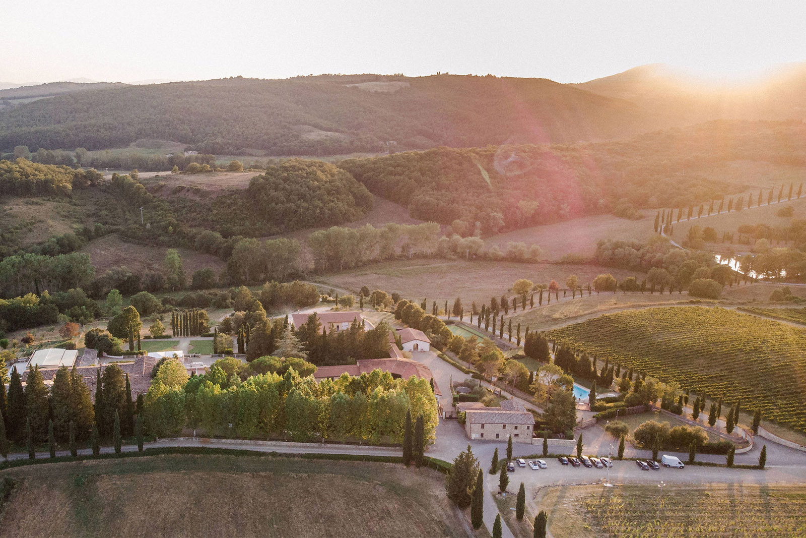 At sunset in the Tuscan countryside, a sweeping aerial view of a villa estate surrounded by vineyards and tree-lined paths is captured in a romantic, editorial landscape style.
