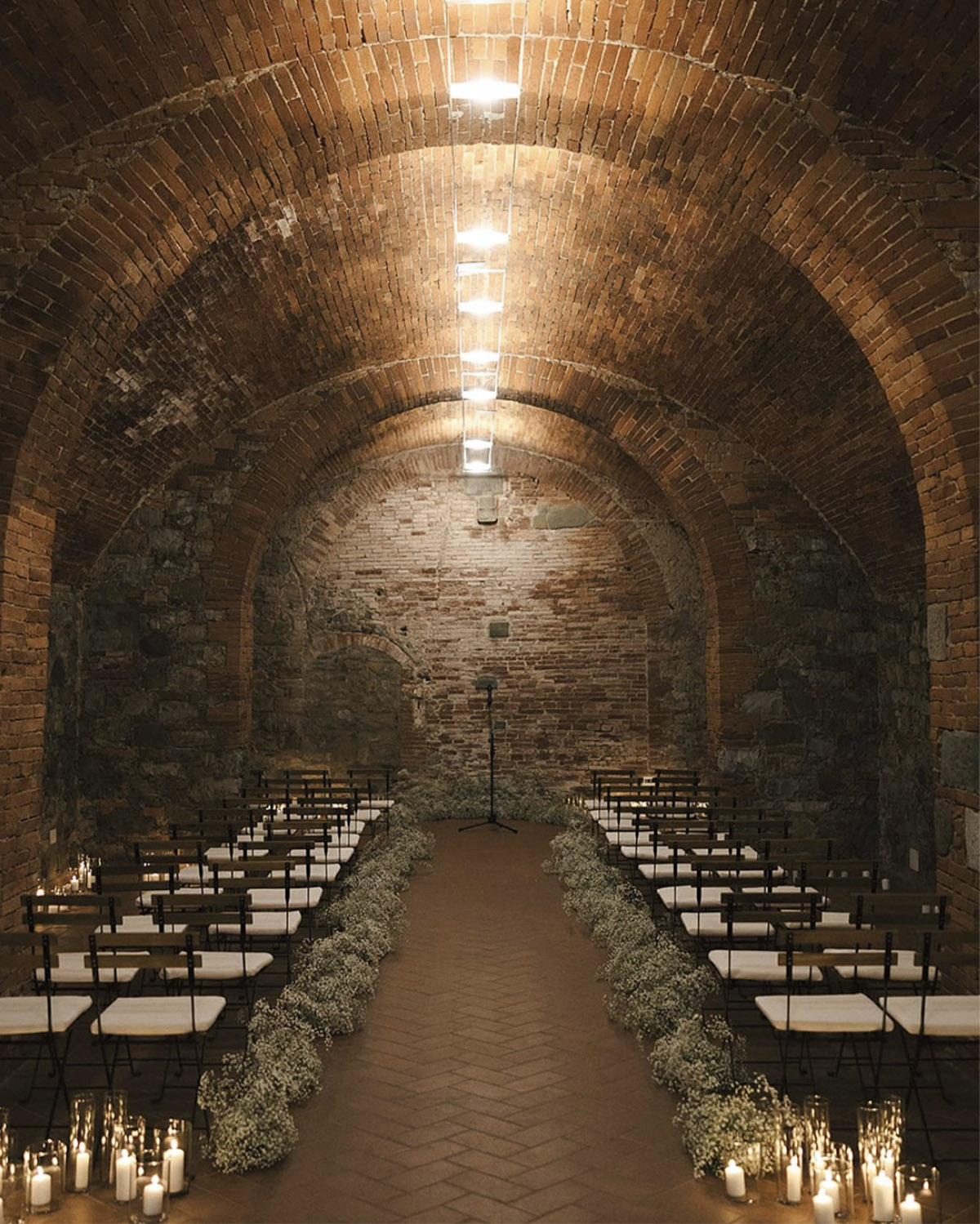 In an arched brick chapel space in Tuscany, rows of minimalist chairs line a candlelit aisle adorned with delicate white florals, captured in a refined and intimate editorial style.
