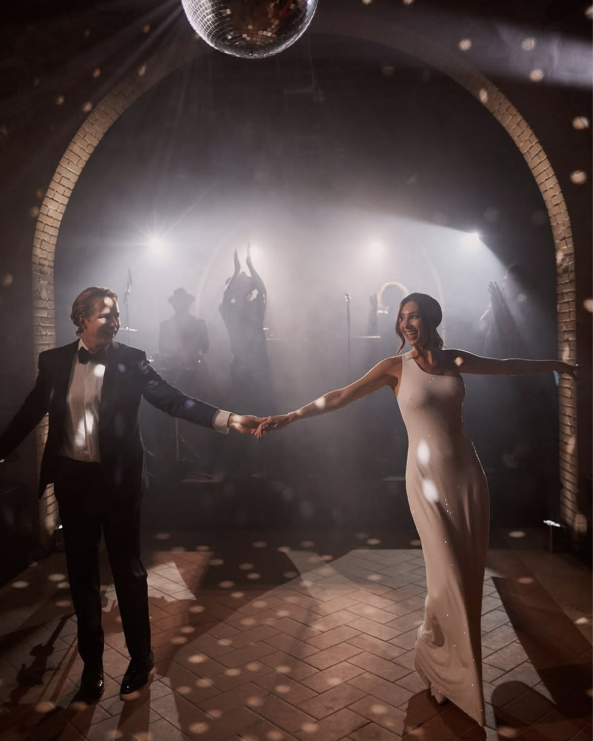 On a softly lit dance floor beneath a disco ball in a Tuscan stone hall, a bride and groom hold hands mid-dance while guests cheer in the background, photographed in a lively, cinematic editorial style.