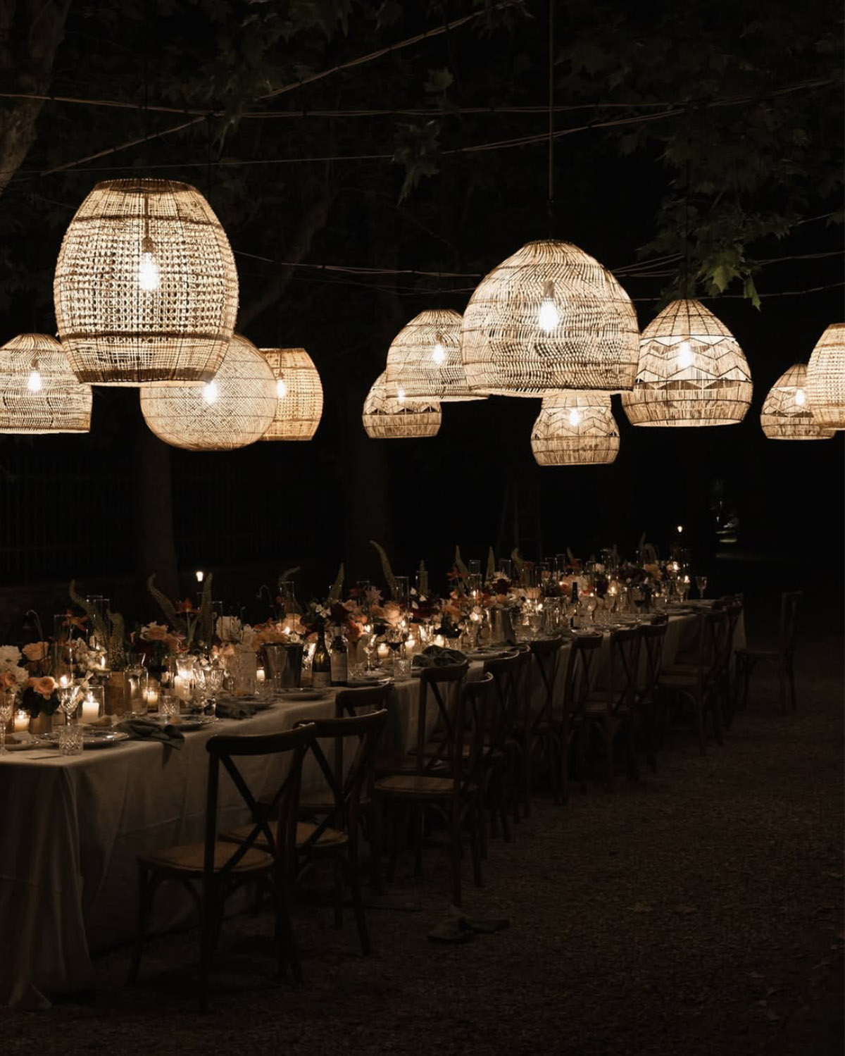 A nighttime outdoor reception beneath woven pendant lights illuminates a long banquet table adorned with candles and florals, styled in a moody, editorial wedding aesthetic.