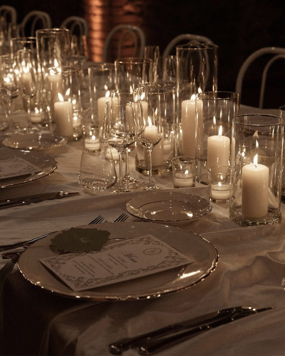 At an elegantly set reception table in a Tuscan venue, crystal glassware and glowing candles surround fine china and printed menus, styled in a luxurious, detail-focused editorial aesthetic.