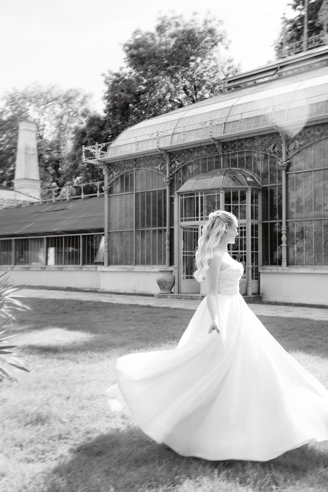Photoshoot in Jevremovac Botanical Garden, Belgrade | Editorial Post-Wedding Photography 19 Belgrade Botanical Garden greenhouse, a bride in a flowing wedding gown twirls across the lawn in front of the historic glasshouse, captured in a dreamy black-and-white editorial style with soft motion and light flare.