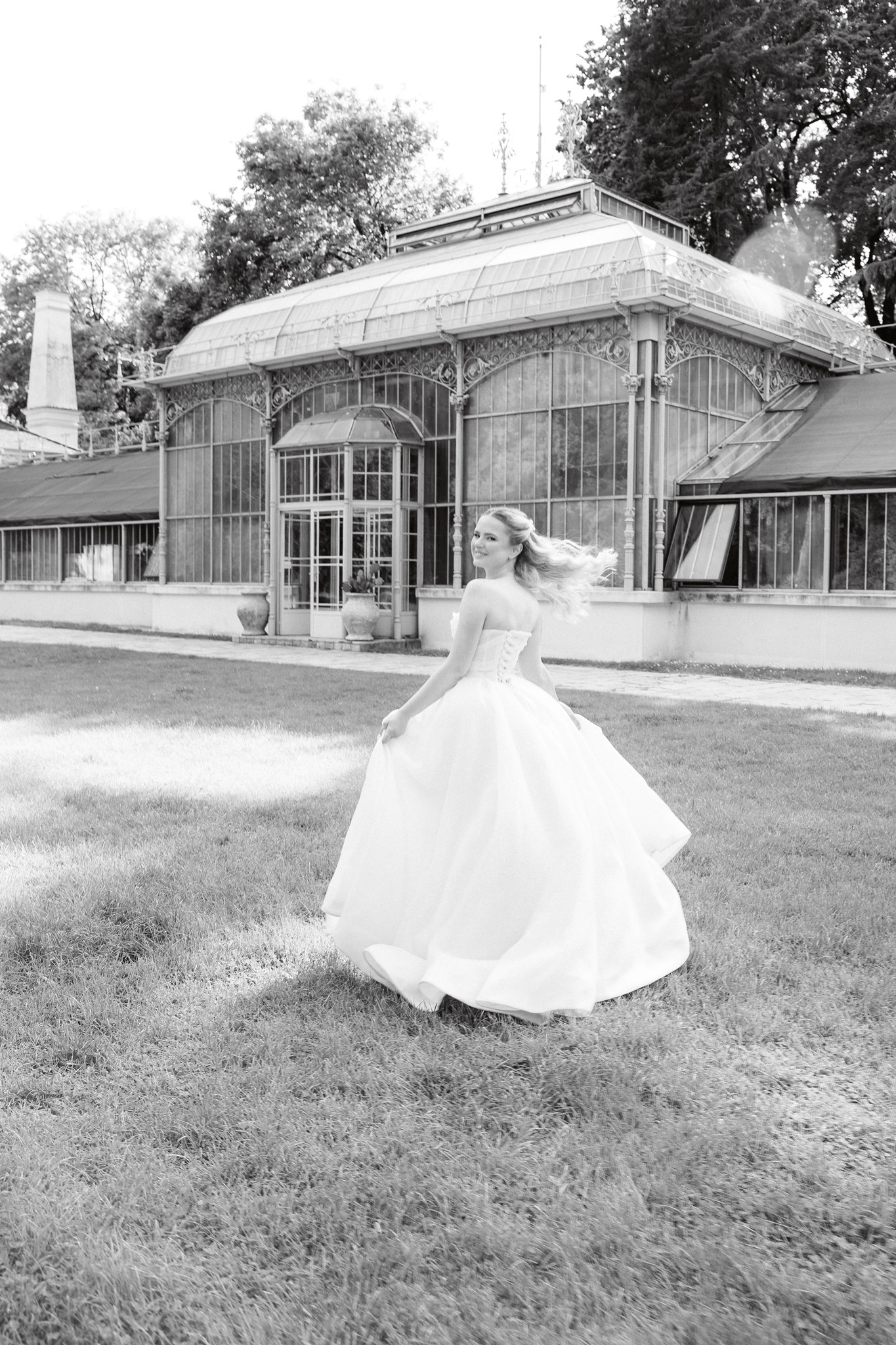 Photoshoot in Jevremovac Botanical Garden, Belgrade | Editorial Post-Wedding Photography 10 Belgrade Botanical Garden greenhouse, a bride spins and smiles over her shoulder while moving across the grass before the ornate conservatory, photographed in a romantic black-and-white editorial style.