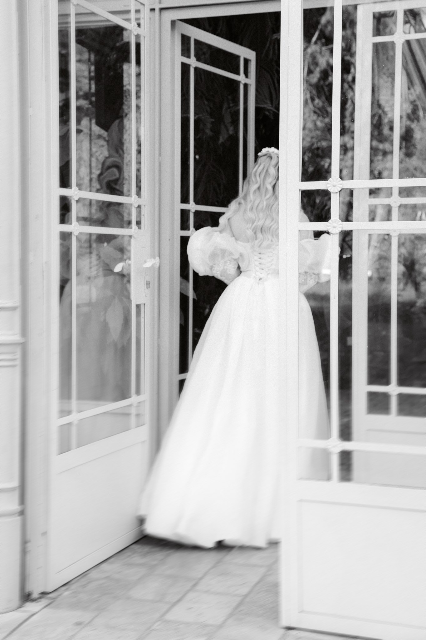 Photoshoot in Jevremovac Botanical Garden, Belgrade | Editorial Post-Wedding Photography 14 Inside the Belgrade Botanical Garden greenhouse entrance, a bride in a full wedding dress walks through open glass doors, captured in a soft-focus black-and-white editorial style.