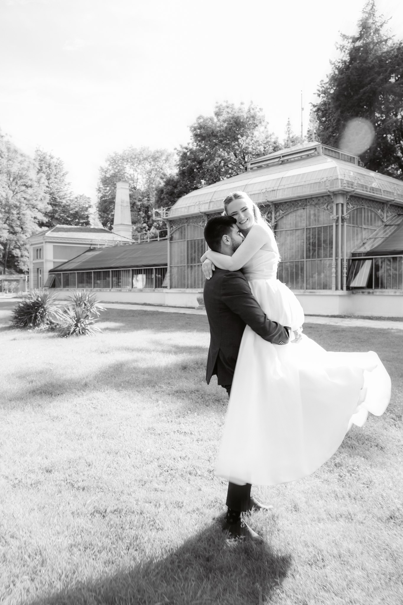 Photoshoot in Jevremovac Botanical Garden, Belgrade | Editorial Post-Wedding Photography 16 Belgrade Botanical Garden greenhouse, a groom lifts his bride in his arms on the lawn outside the conservatory, photographed in a timeless black-and-white editorial style with natural light.