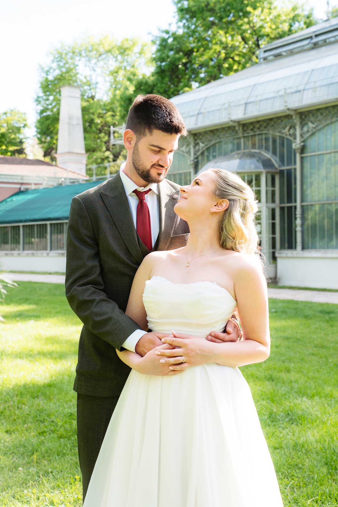 Photoshoot in Jevremovac Botanical Garden, Belgrade | Editorial Post-Wedding Photography 20 Belgrade Botanical Garden greenhouse, a bride and groom stand embracing and gazing at one another on the lawn beside the conservatory, photographed in a bright and elegant editorial wedding style.