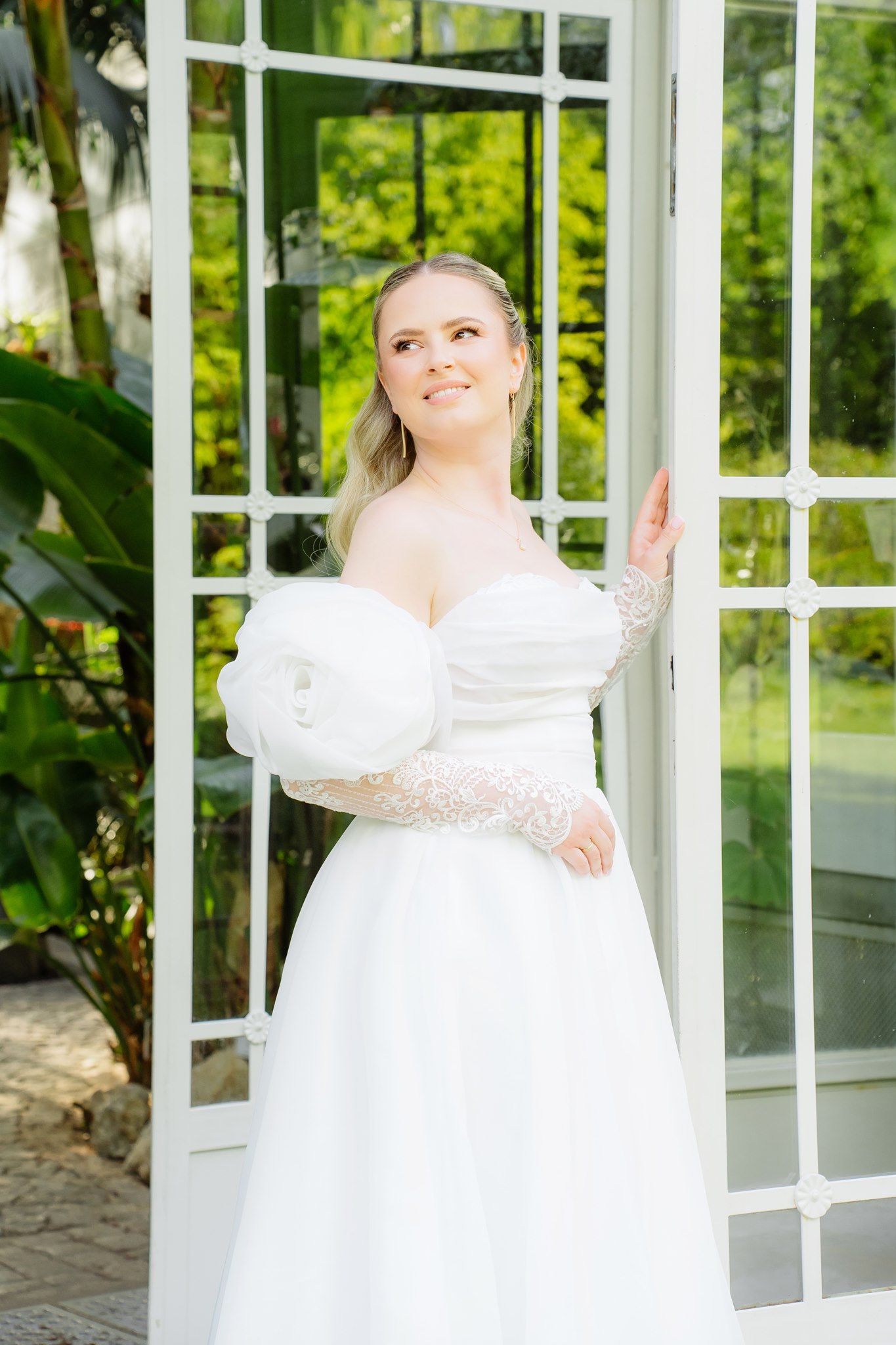 Photoshoot in Jevremovac Botanical Garden, Belgrade | Editorial Post-Wedding Photography 13 Belgrade Botanical Garden greenhouse entrance, a bride in an off-the-shoulder wedding gown poses beside open glass doors surrounded by lush greenery, photographed in a refined editorial bridal style.