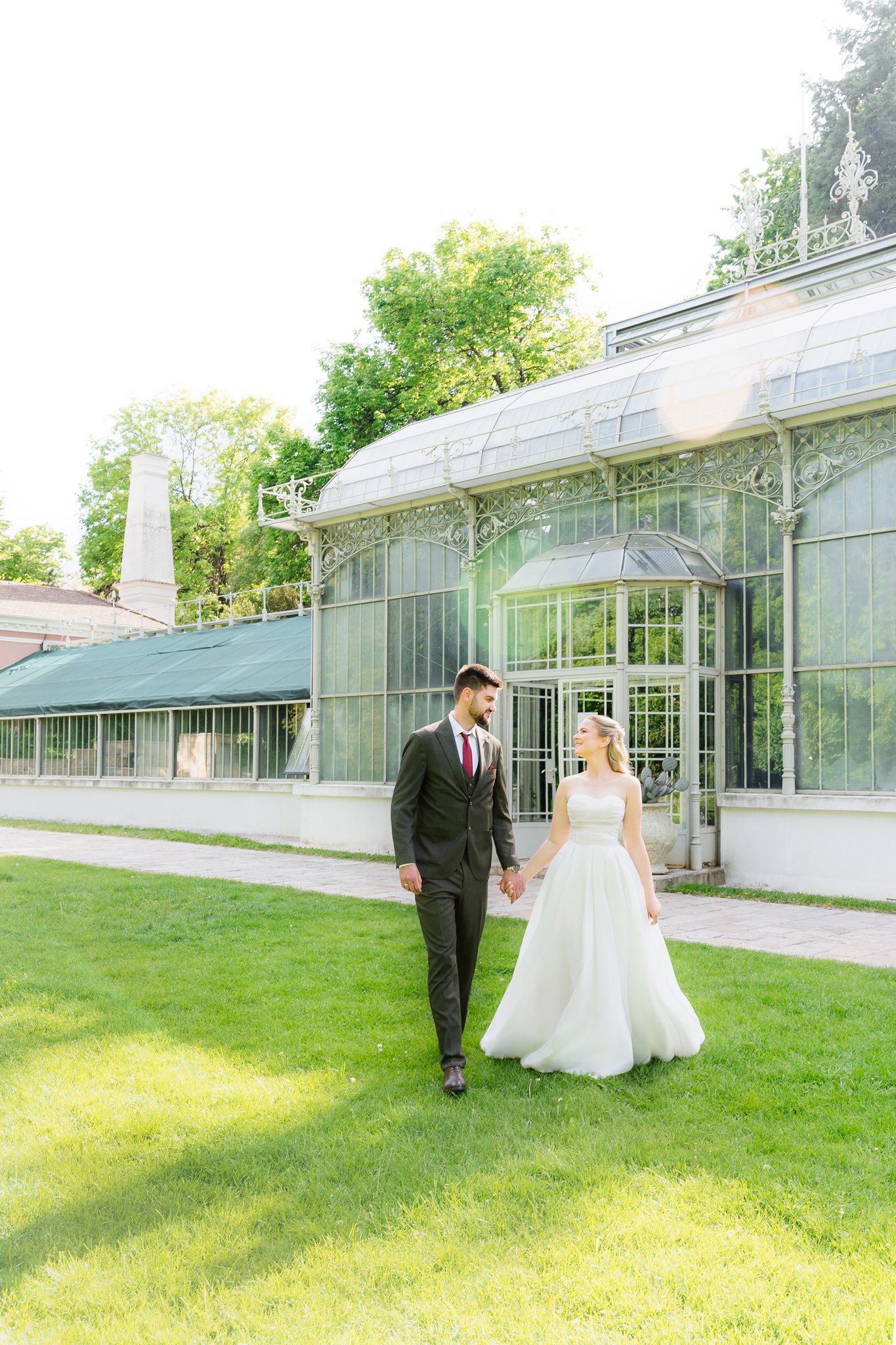 MIJO WEDDINGS 20 Belgrade Botanical Garden greenhouse, a bride and groom walk hand in hand across the lawn in front of the historic conservatory, captured in a light-filled editorial wedding style.