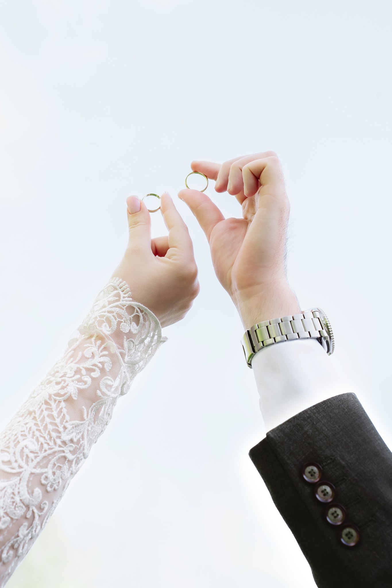 Photoshoot in Jevremovac Botanical Garden, Belgrade | Editorial Post-Wedding Photography 11 Low-angle shot of a bride and groom's hands raised against a pale sky, each holding a gold wedding band between their fingers — her lace sleeve and his dark suit cuff visible.