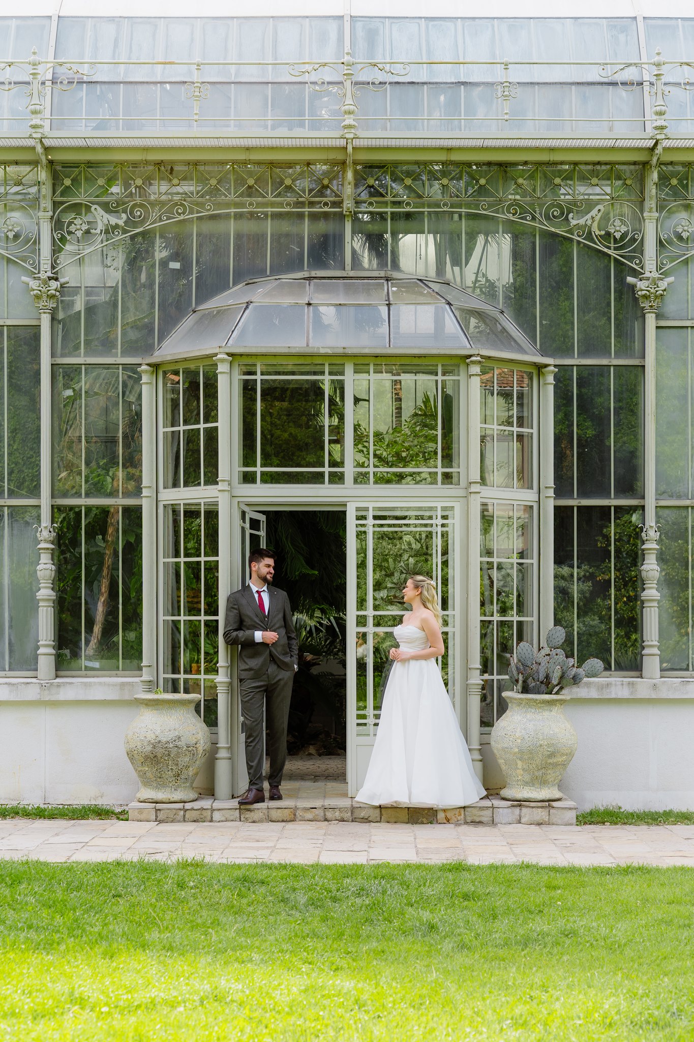 Bride and groom standing at opposite sides of the ornate iron-and-glass entrance of the Belgrade Botanical Garden greenhouse, smiling across at each other in afternoon light.