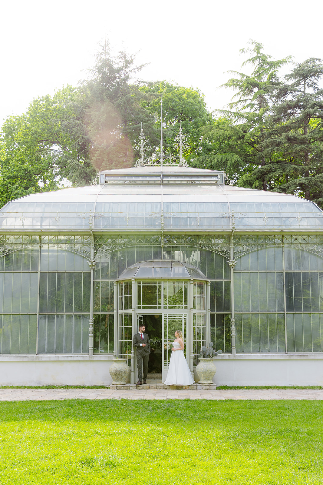 MIJO WEDDINGS 5 Editorial photo of the couple standing in the arched doorway of the Jevremovac greenhouse. Ornate Victorian glass-and-iron structure surrounded by mature trees.