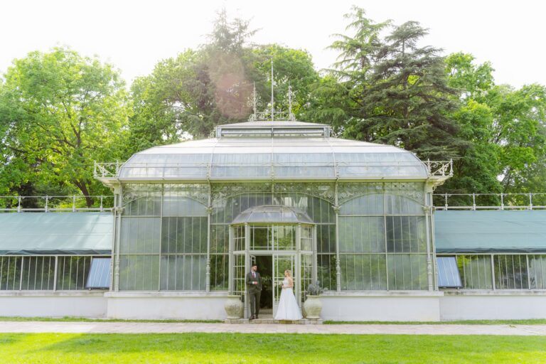 Photoshoot in Jevremovac Botanical Garden, Belgrade | Editorial Post-Wedding Photography 1 Wide-angle editorial of the couple standing in the arched doorway of the full Jevremovac greenhouse facade, dwarfed by the ornate Victorian glass-and-iron structure surrounded by mature trees.