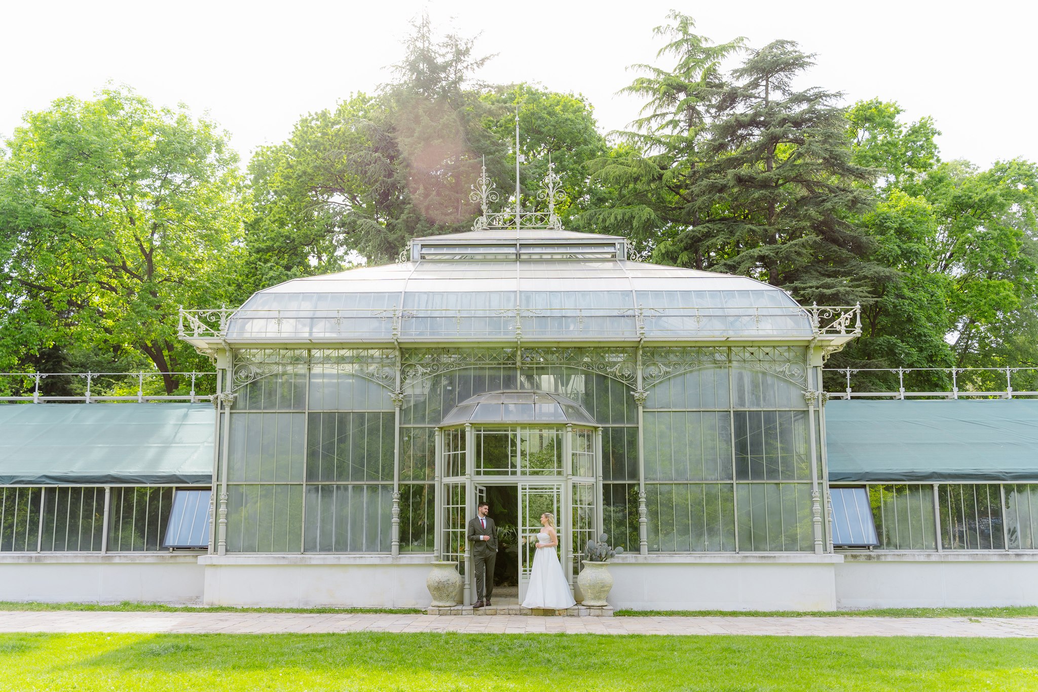 Wide-angle editorial of the couple standing in the arched doorway of the full Jevremovac greenhouse facade, dwarfed by the ornate Victorian glass-and-iron structure surrounded by mature trees.