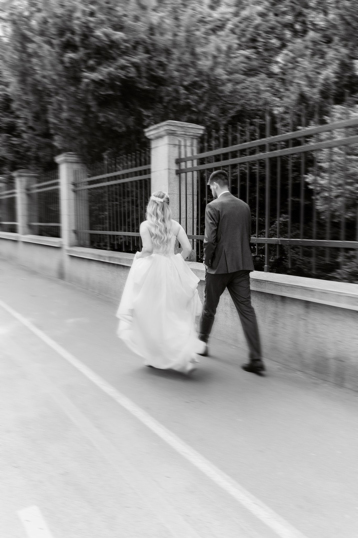 Photoshoot in Jevremovac Botanical Garden, Belgrade | Editorial Post-Wedding Photography 12 Intentionally motion-blurred black and white shot of the bride and groom walking hand in hand along a Belgrade street beside an iron fence, conveying movement and spontaneity.