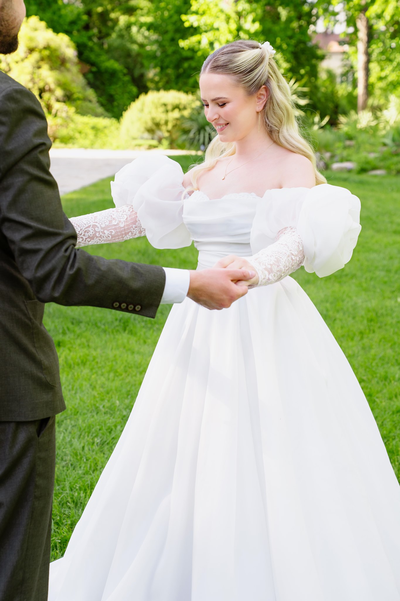 Photoshoot in Jevremovac Botanical Garden, Belgrade | Editorial Post-Wedding Photography 15 Candid editorial of the bride laughing and holding the groom's hands on the botanical garden lawn, her voluminous off-shoulder gown with dramatic puff sleeves catching the sunlight.