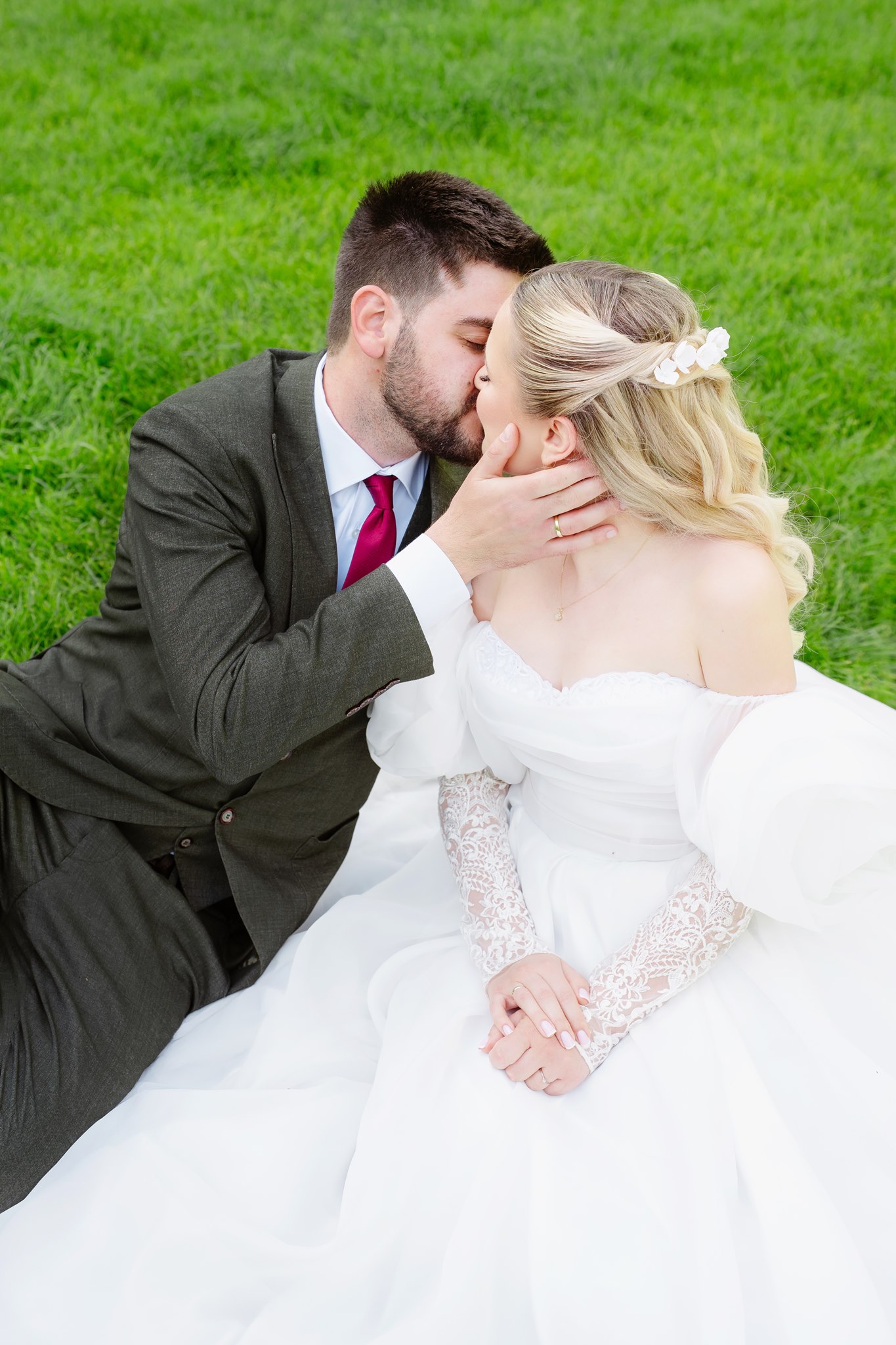Bride and groom sitting on a lush green lawn, the groom kissing her on the cheek while she holds his hand, her voluminous lace-sleeved gown spread around them.