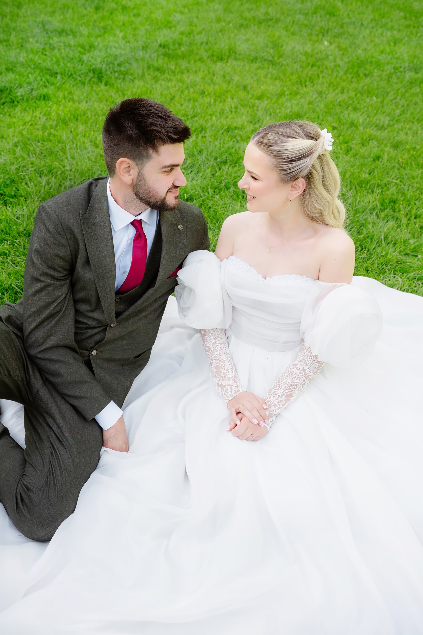 Photoshoot in Jevremovac Botanical Garden, Belgrade | Editorial Post-Wedding Photography 9 Bride and groom seated together on a bright green lawn, gazing at each other and holding hands, her billowing tulle gown and his burgundy tie catching the daylight.