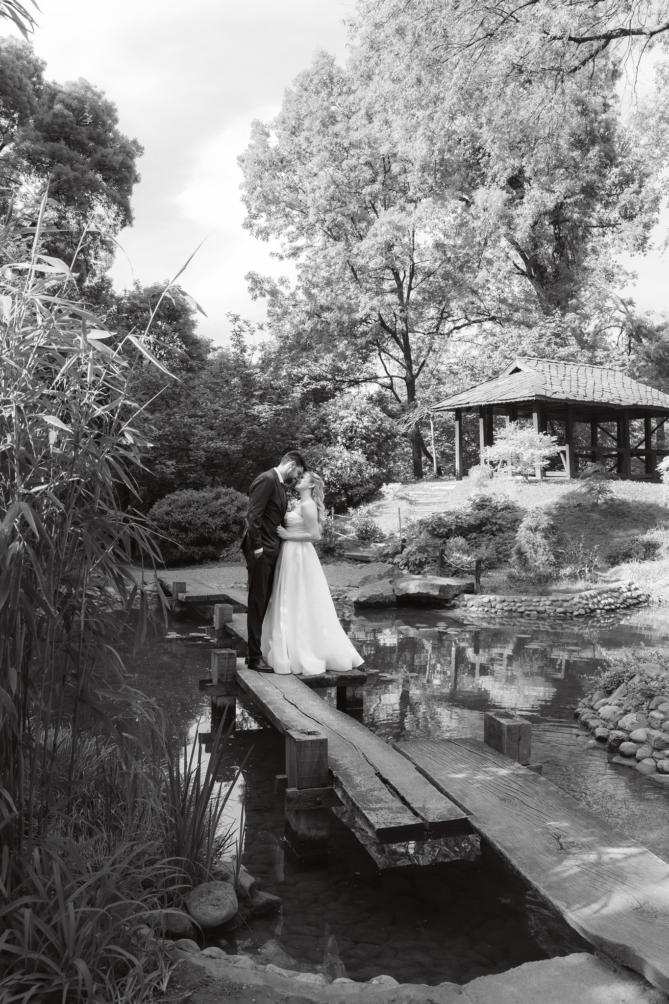MIJO WEDDINGS 3 Black and white editorial of a bride and groom embracing on a rustic wooden bridge over a koi pond in the Japanese Garden, a traditional pavilion visible in the background.
