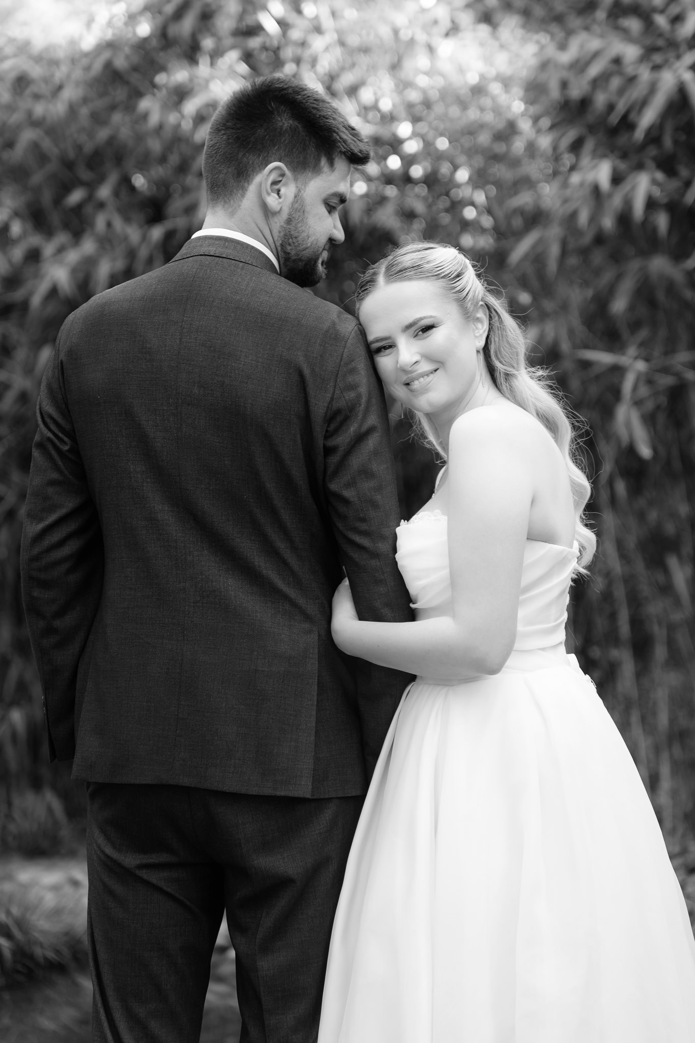 Black and white editorial portrait of a bride resting her head on the groom's shoulder in a bamboo grove, smiling softly at the camera while he gazes down at her.