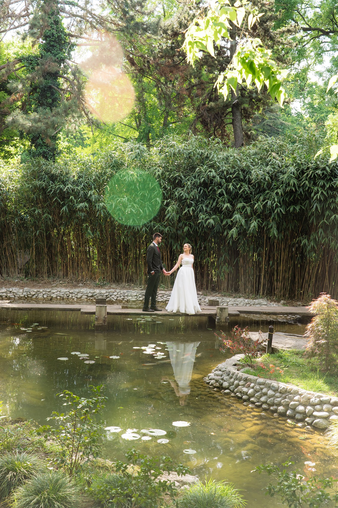 MIJO WEDDINGS 18 Dreamy editorial of the bride and groom holding hands on a dock above a lily pad pond, their reflection mirrored below, flanked by tall bamboo and sun flare.