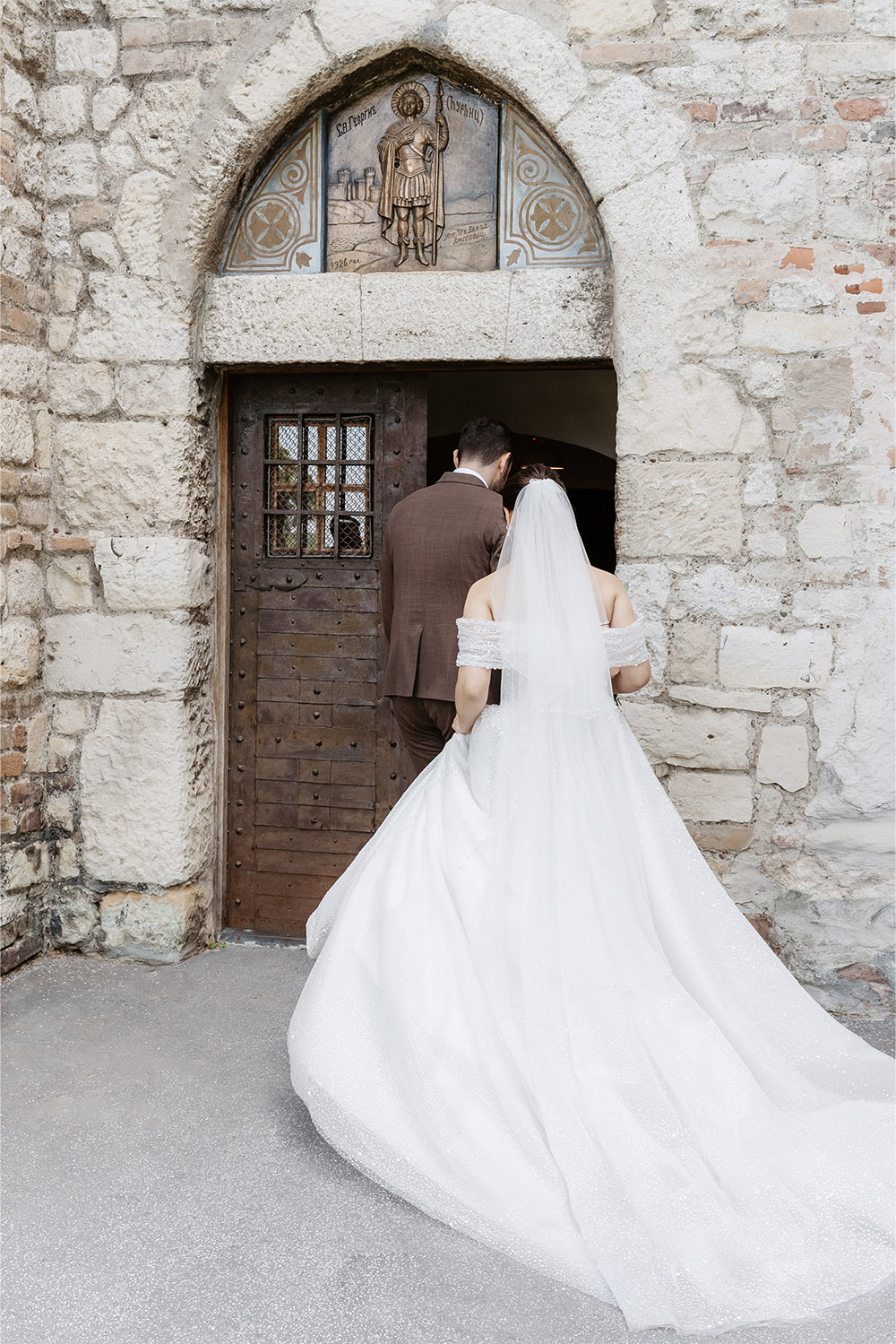 At Ružica Church within Belgrade’s Kalemegdan Fortress, a bride and groom enter the historic stone chapel in an editorial wedding photograph that highlights the bride’s flowing gown and veil against the medieval architecture.