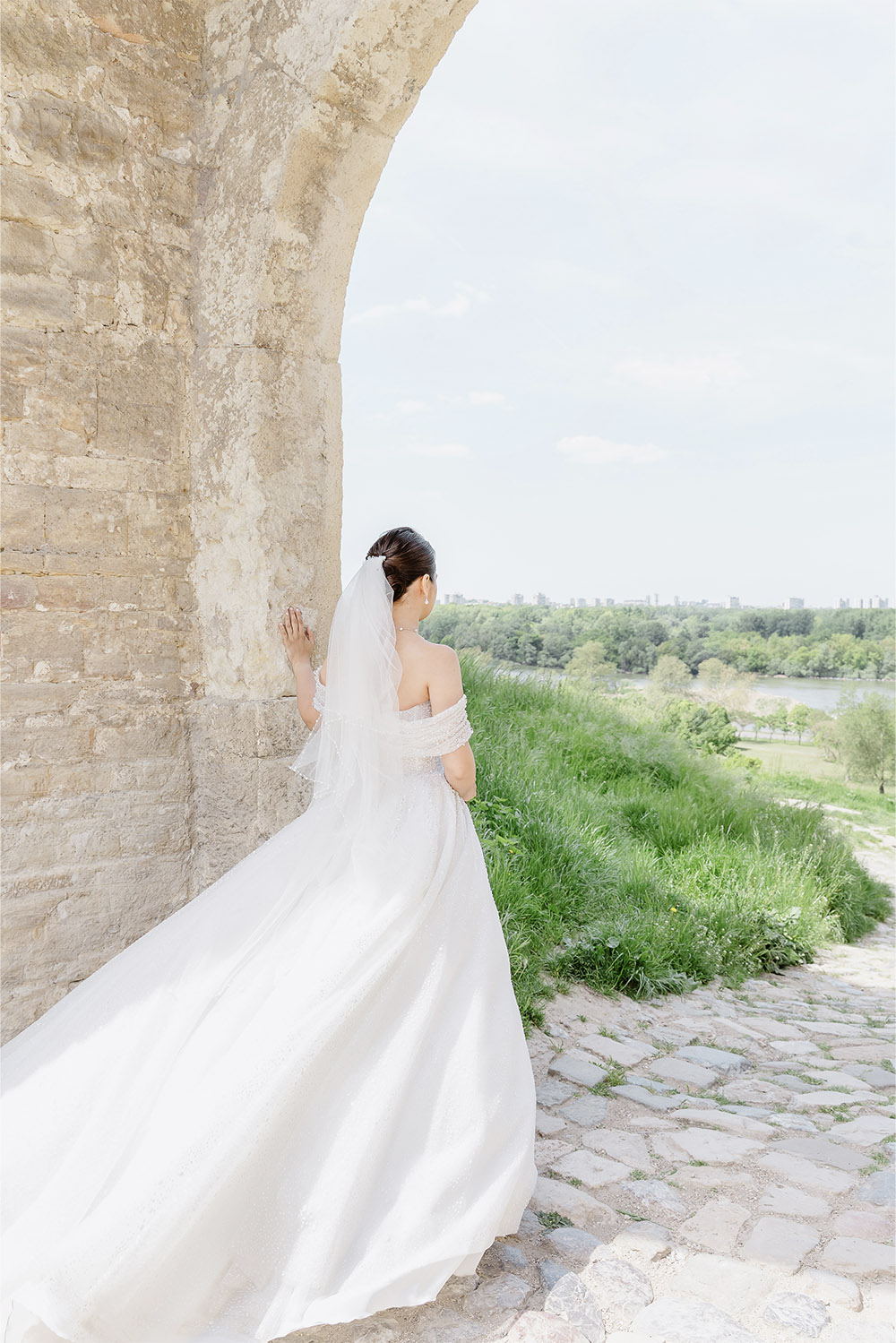 At Kalemegdan Fortress in Belgrade, the bride stands beneath a historic stone arch with her gown flowing across the cobblestones in a romantic editorial wedding portrait.