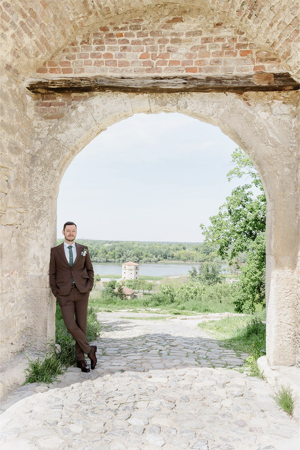 At Kalemegdan Fortress in Belgrade, the groom leans casually beneath a weathered stone arch overlooking the river in a refined editorial wedding portrait.