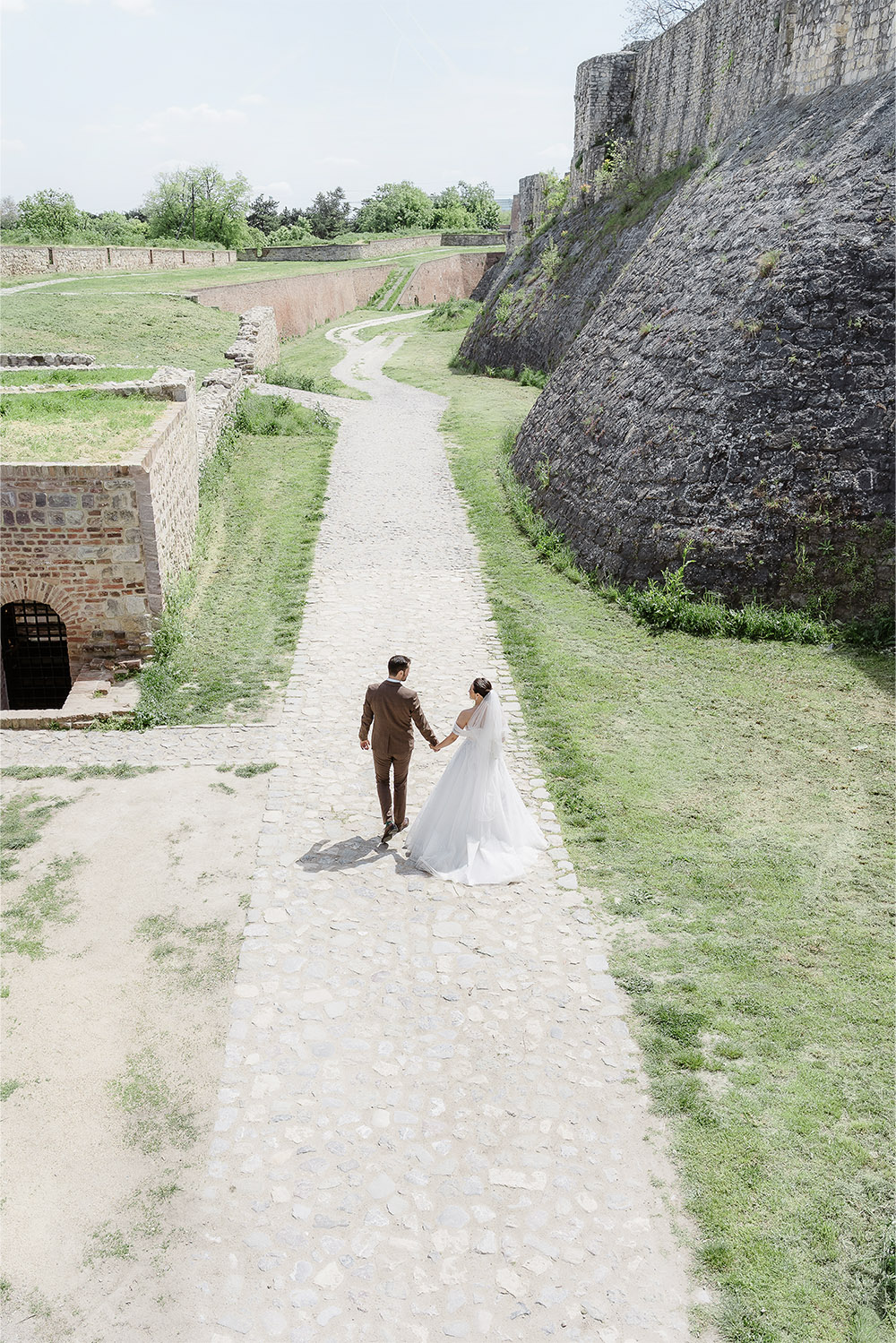 At Kalemegdan Fortress in Belgrade, the bride and groom walk hand in hand along a cobblestone path framed by ancient stone walls in an elegant editorial wedding photograph.