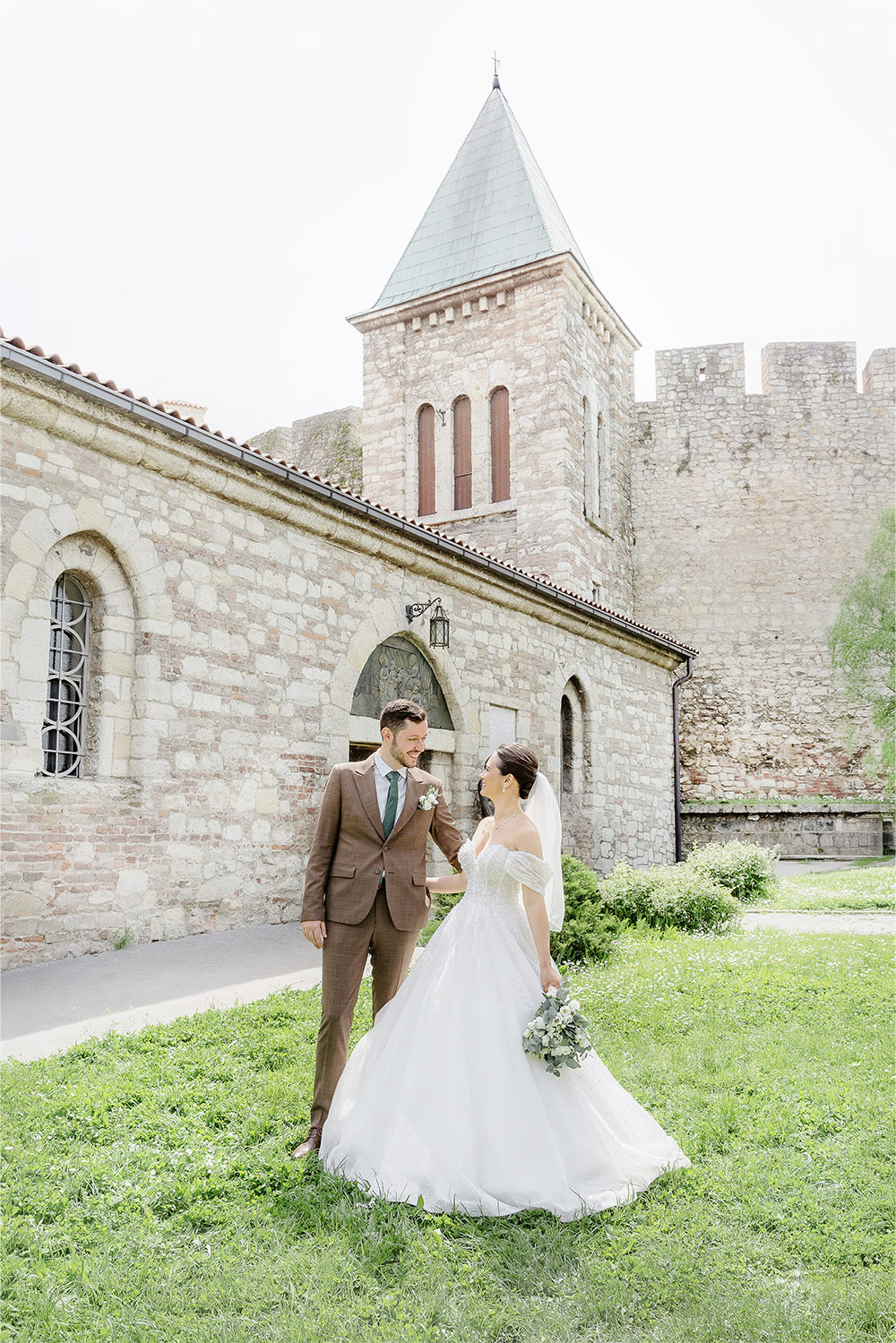 Outside Ružica Church at Kalemegdan Fortress in Belgrade, a bride and groom share a joyful moment on the lawn in a light-filled editorial wedding portrait framed by the church and fortress walls.