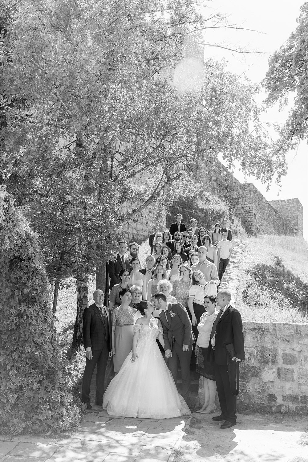At Kalemegdan Fortress in Belgrade, a bride and groom pose with family and guests beneath leafy trees and stone ramparts in a classic black-and-white editorial wedding group portrait.