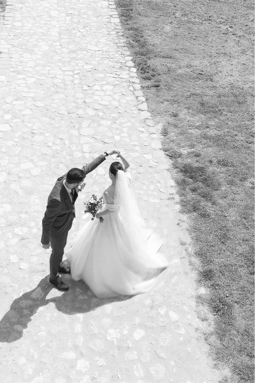 At Kalemegdan Fortress in Belgrade, the bride twirls beneath her partner’s raised hand along a cobblestone path in a dramatic black-and-white editorial wedding photograph.