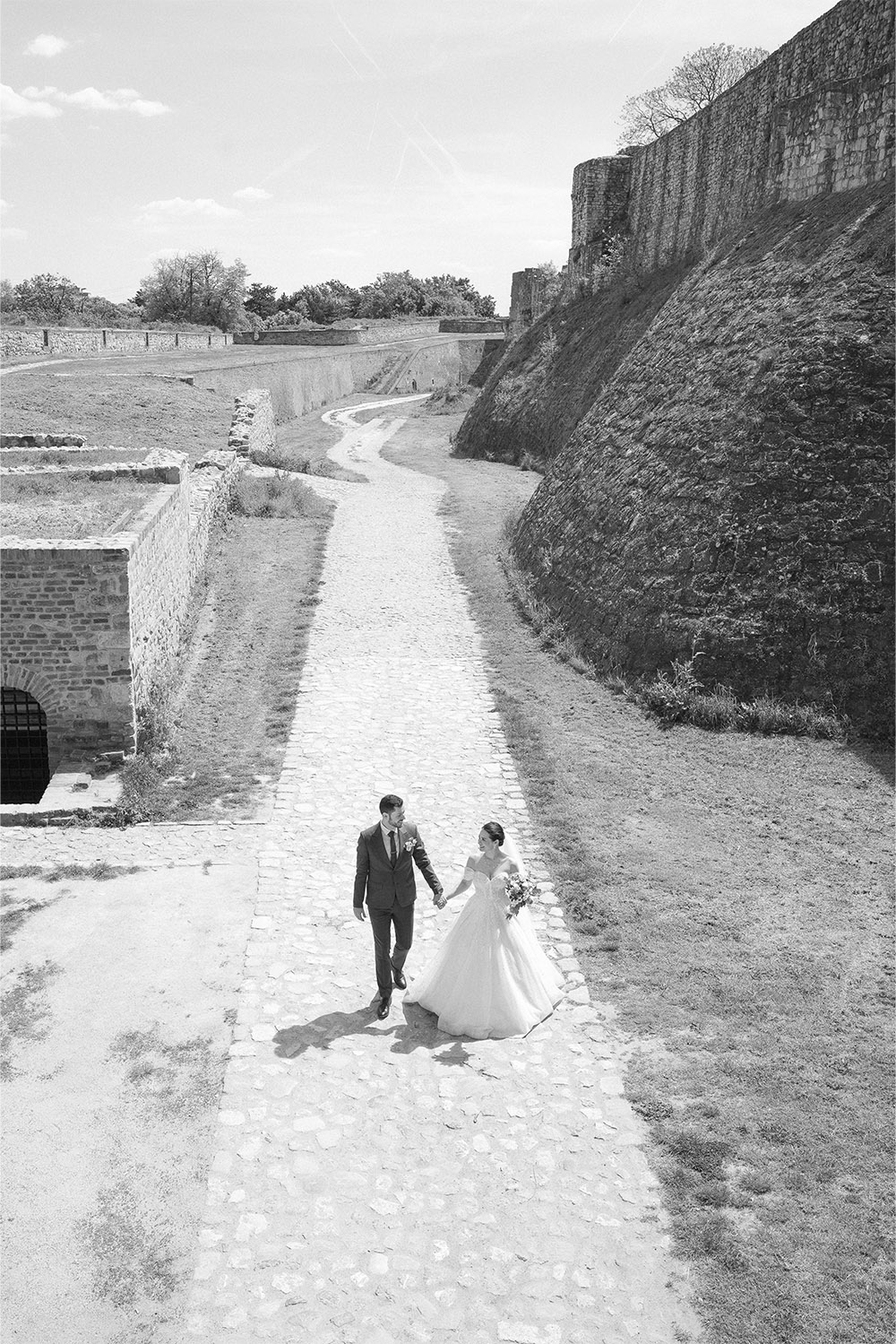 At Kalemegdan Fortress in Belgrade, the bride and groom walk together beneath towering stone walls in a black-and-white editorial wedding portrait captured from above.