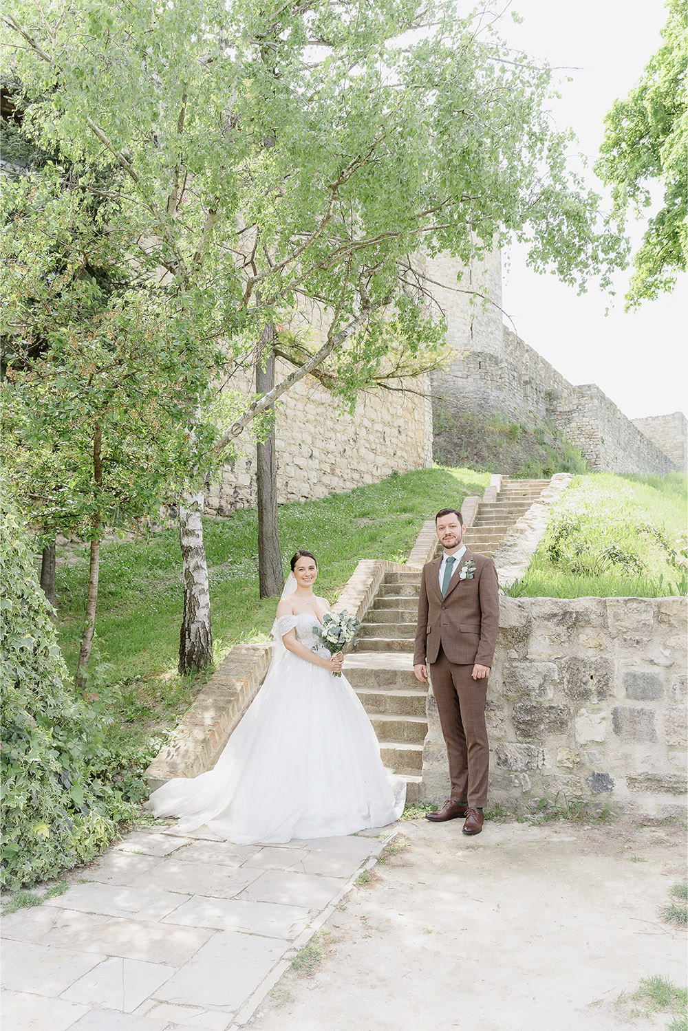 At Kalemegdan Fortress in Belgrade, the bride and groom stand beside stone steps and leafy trees within the historic grounds in a bright editorial wedding portrait.
