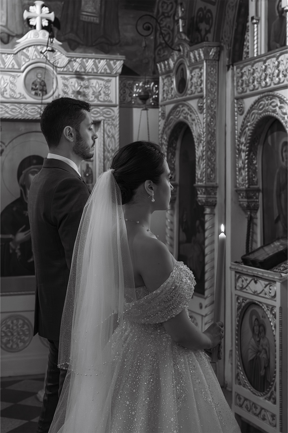 Inside Ružica Church in Belgrade, a bride and groom stand together holding ceremonial candles during their Orthodox wedding in an intimate black-and-white editorial photograph.