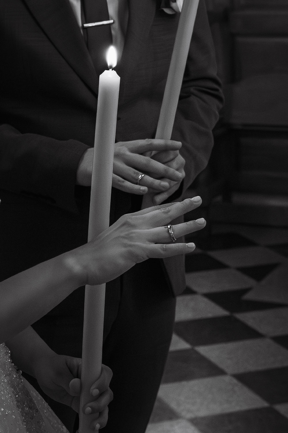 During an Orthodox wedding ceremony at Ružica Church in Belgrade, a close-up editorial black-and-white photograph captures the bride and groom holding lit ceremonial candles and displaying their wedding rings.
