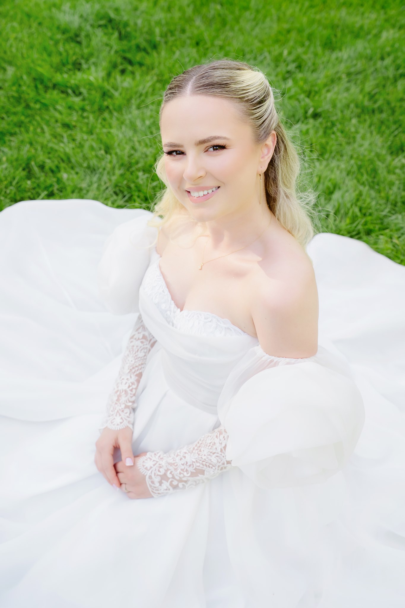 Close-up editorial portrait of the bride seated on green grass, smiling warmly at the camera, her off-shoulder gown with lace sleeves fanning out around her.