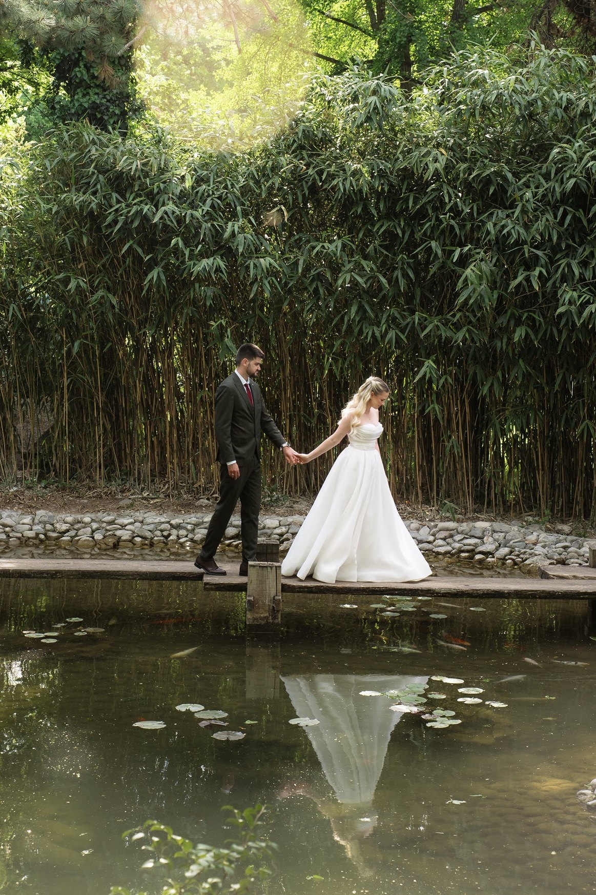 Bride and groom walking hand in hand along a wooden dock beside a lily pad pond, their reflections visible in the still water, a dense bamboo grove behind them bathed in dappled light.
