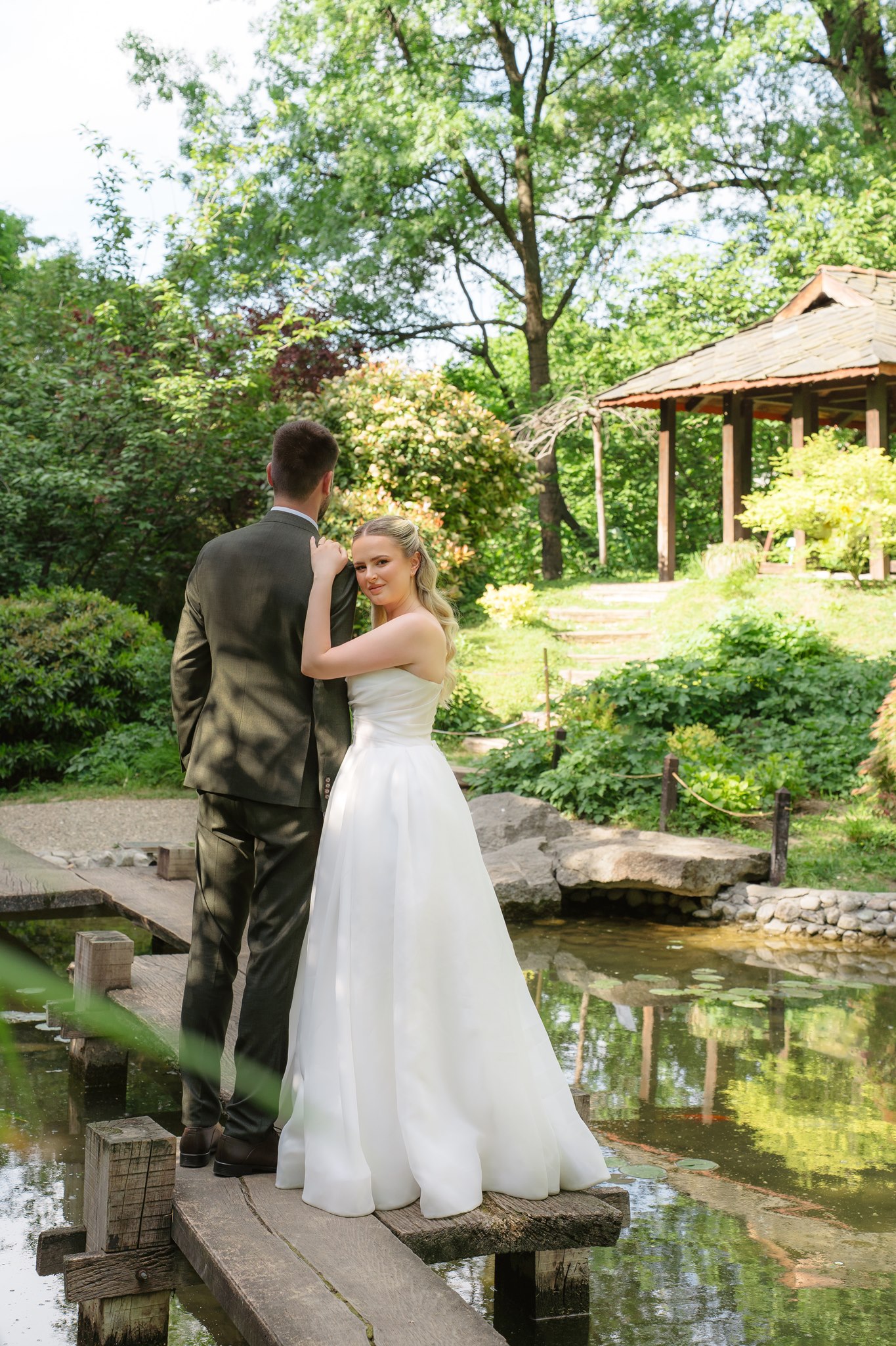 Bride glancing back at the camera over her shoulder on a wooden bridge in the Japanese Garden, the groom facing the koi pond and a tiled gazebo beyond.
