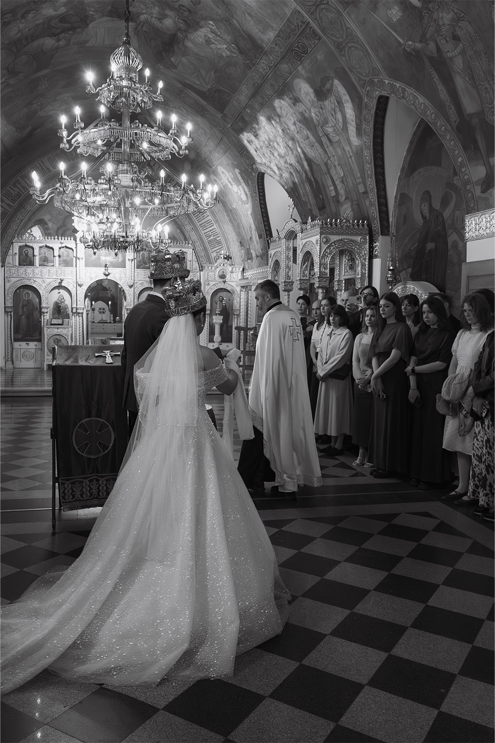 During an Orthodox wedding ceremony at Ružica Church in Belgrade, a crowned bride and groom stand before the priest and their guests in an elegant black-and-white editorial photograph.