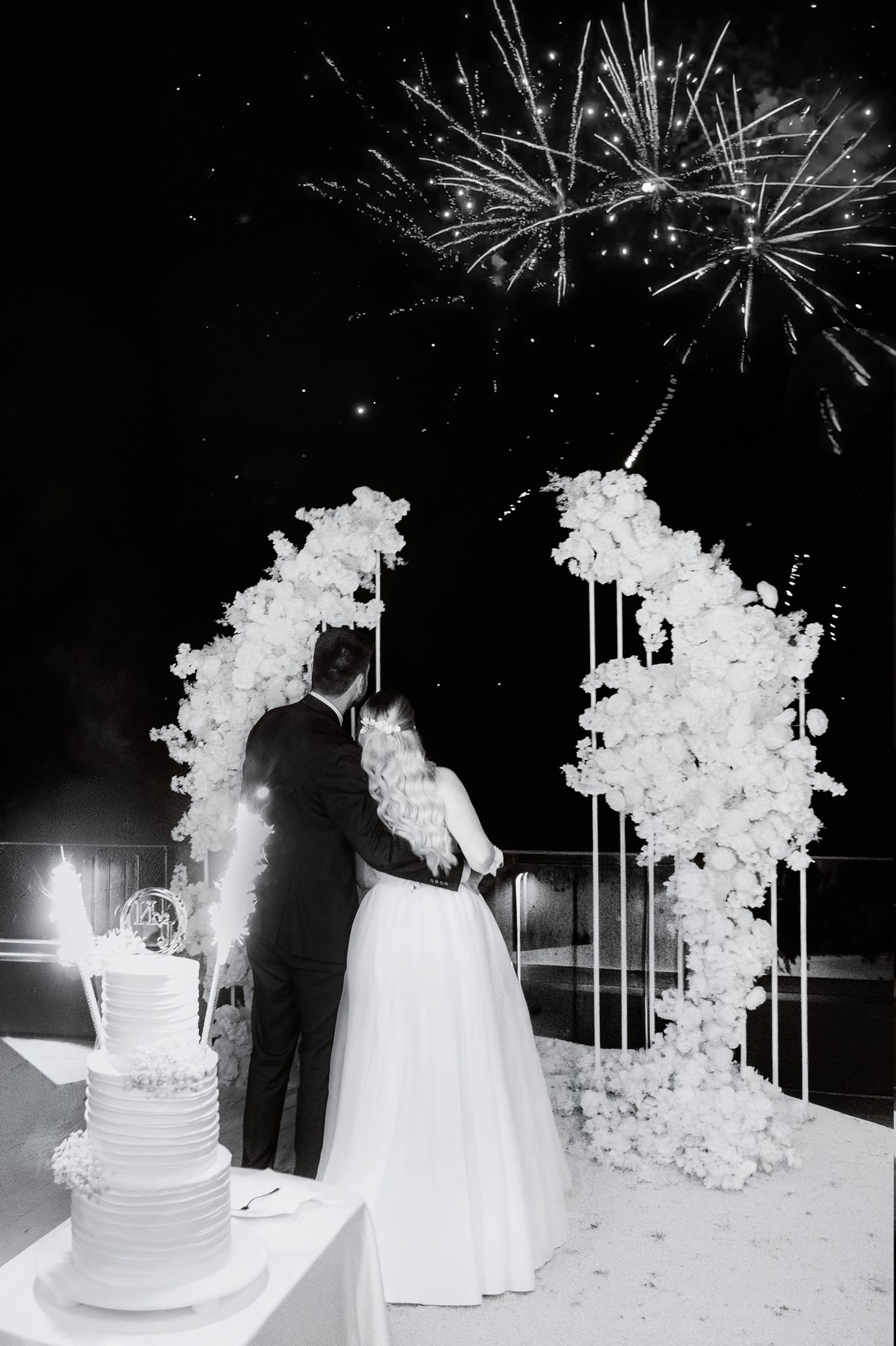 MIJO WEDDINGS 27 At an outdoor wedding reception beneath a night sky lit by fireworks, a newly married couple stands arm in arm beside their cake in an elegant editorial-style photograph filled with celebration and drama.