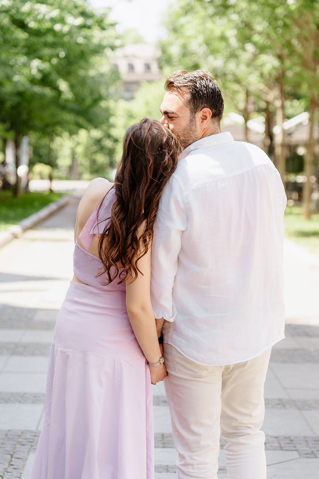 Editorial engagement photo of a couple embracing while walking through a leafy boulevard in central Belgrade during golden summer light.