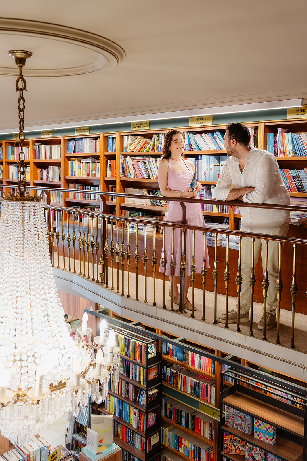 Editorial engagement portrait of a couple smiling and talking on the gallery level of Akademija Bookstore in Belgrade with books lining the walls.