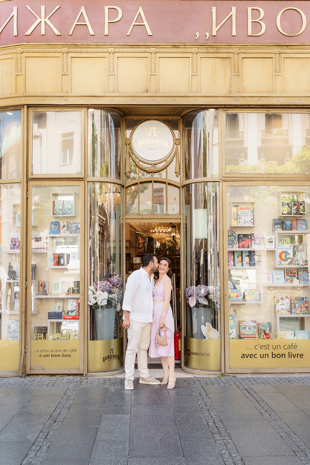 Editorial engagement photo of a couple standing in front of Akademija Bookstore in Belgrade as they share a joyful moment outside the historic entrance.