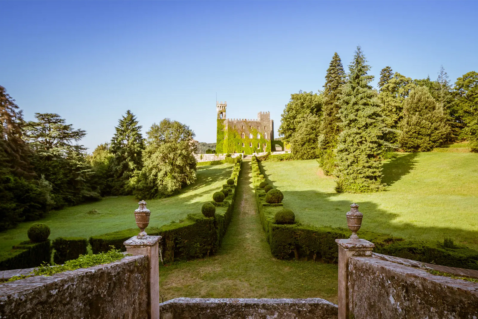 An editorial landscape photograph of Castello di Celsa in Tuscany, Italy, with a tree-lined pathway leading through manicured gardens toward the ivy-covered castle.