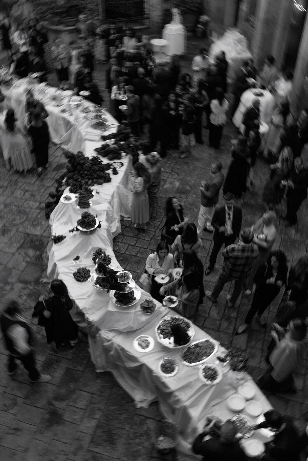 An editorial overhead photograph of an outdoor wedding dinner at Castello di Celsa in Tuscany, Italy, with guests gathered around an elegant buffet in the castle courtyard.