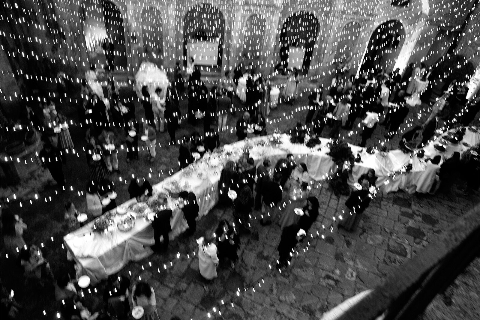 An editorial photograph of an evening wedding reception at Castello di Celsa in Tuscany, Italy, with guests dining beneath glowing string lights in the historic courtyard.