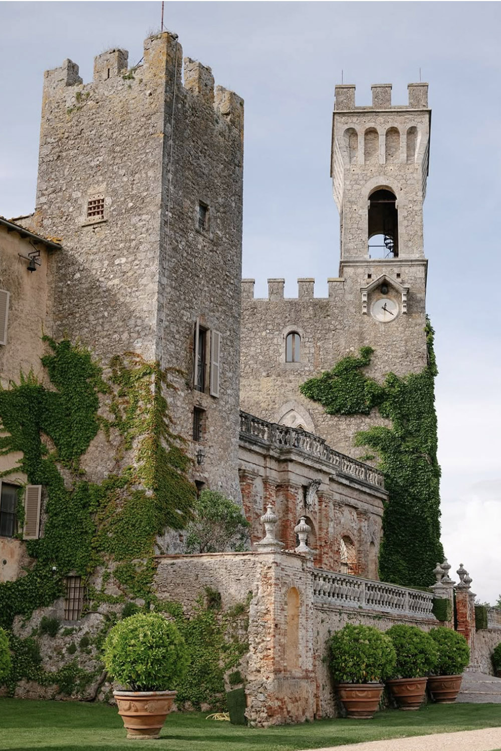 An editorial photograph of Castello di Celsa in Tuscany, Italy, showcasing the ivy-covered medieval castle towers and historic stone architecture against a soft sky.