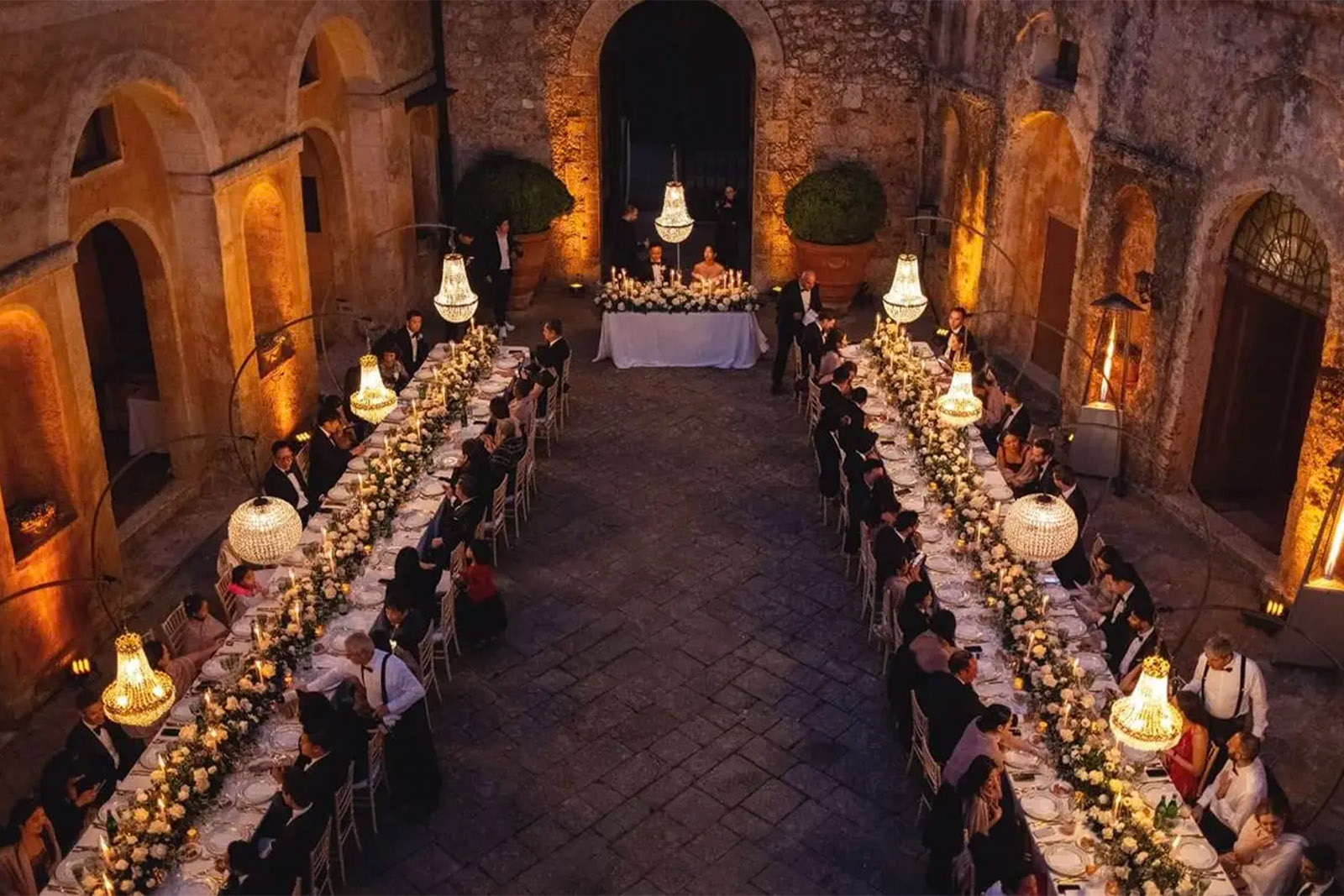 An editorial photograph of an outdoor wedding reception at Castello di Celsa in Tuscany, Italy, with candlelit banquet tables set in the historic stone courtyard.