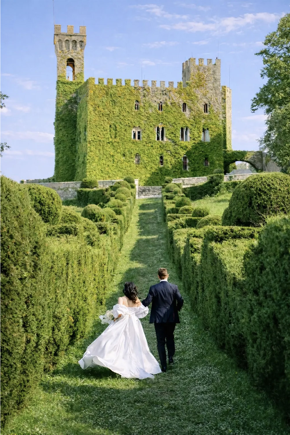 An editorial photograph of Castello di Celsa in Tuscany, Italy, with a bride and groom walking hand in hand through the formal gardens toward the ivy-covered castle.