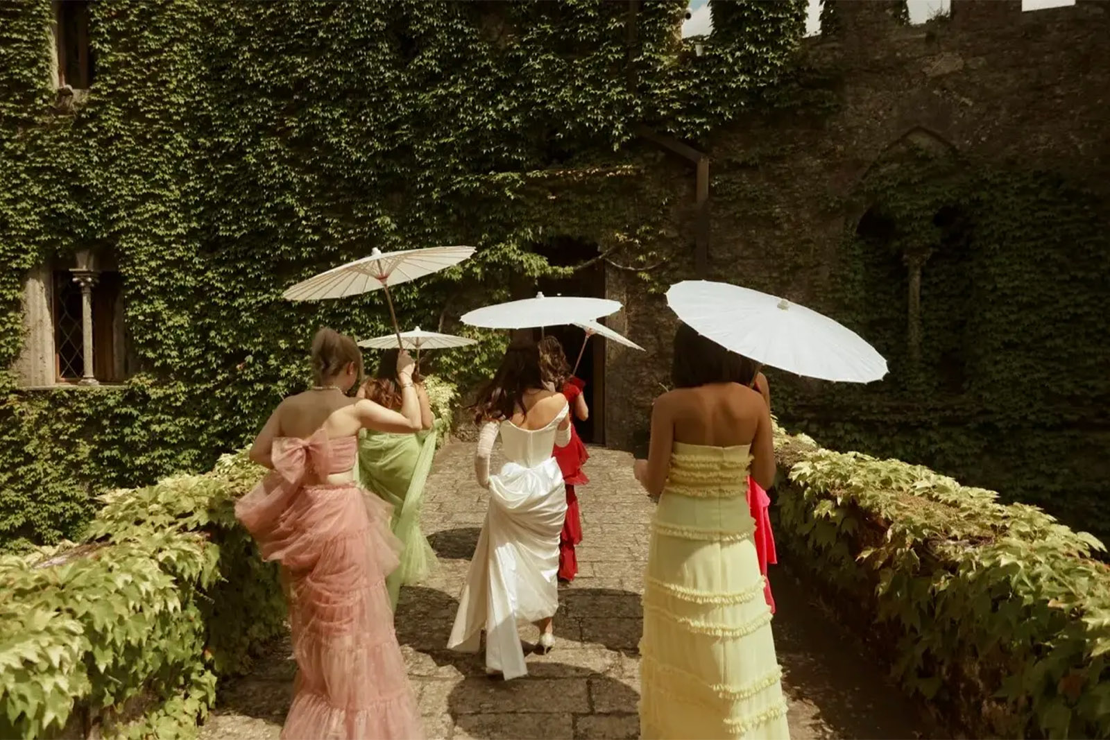 An editorial photograph of wedding guests walking through the ivy-covered courtyard of Castello di Celsa in Tuscany, Italy, carrying parasols on a sunny afternoon.